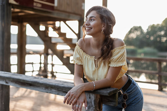 Pretty happy girl with a smile in fashionable denim clothes with summer yellow top stands on a wooden pier near the lake