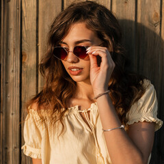 Beautiful model woman hipster put on a fashion sunglasses near vintage wooden wall at sunset