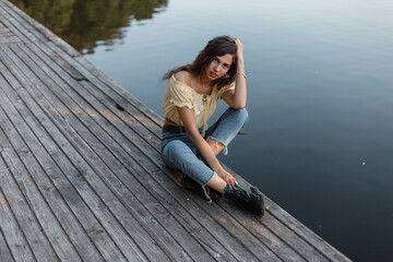 Fototapeta na wymiar Fashionable beauty woman with curly hair in blue jeans with yellow top blouse and boots sits and rest on a vintage wooden pier near lake