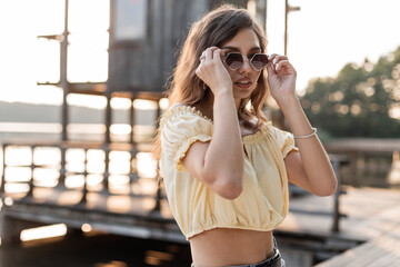 Cool hipster young pretty woman in fashion yellow blouse puts up stylish round vintage sunglasses outdoors on pier by the lake