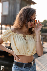 Fashion pretty young woman hipster in a stylish yellow blouse with blue jeans and trendy sunglasses on the pier near the beach.