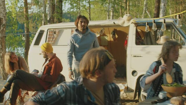 Young friends cooking food, chatting and playing guitar at campsite in forest while preparing for outdoor party in nature