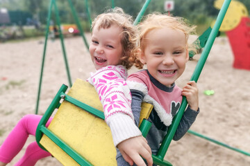 Two little blonde caucasian girls swing on a swing.Children look at the camera and laugh heartily.Children's emotions.