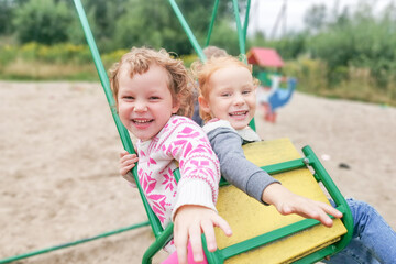Two little blonde caucasian girls swing on a swing.Children look at the camera and laugh heartily.Children's emotions.