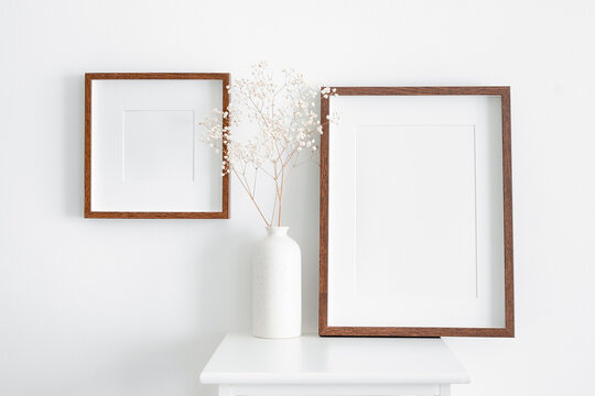 Stylish and modern frames mockup for artwork, print or photo presentation. Blank wooden frames on white wall with furniture and ceramic vase with dry gypsophila plant.