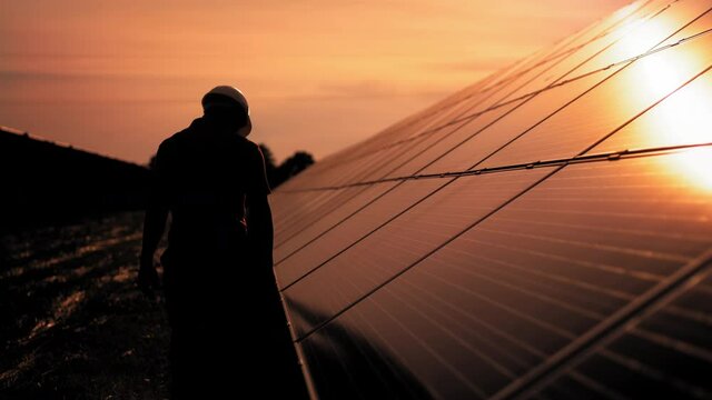 Assistance technical worker in uniform is checking an operation and efficiency performance of photovoltaic solar panels. Unidentified solar power engineer touches solar panels with his hand at sunset