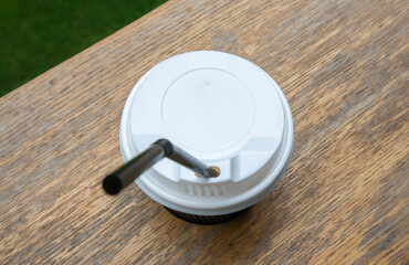 One black paper coffee cup with lid outdoors in summer in sunny weather on a wooden table of a cafe, coffee shop or restaurant.