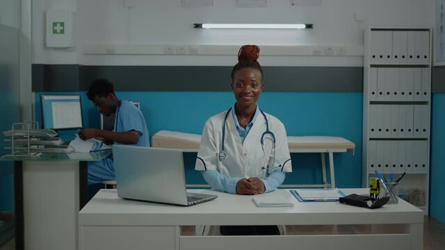 Portrait of african american woman with doctor profession wearing white coat and stethoscope at medical facility. Black medic smiling looking at camera in cabinet with nurse in background