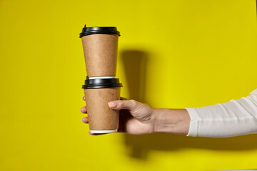 Hands holding two cups brown paper with black lid. Two coffee special offer or promo. Hands holding two cups on yellow screen background. Tea or Coffee to go. Brown paper cup with black lid.