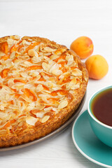 Closeup apricot pie decorated with almond petals and cup of tea on white wooden table.