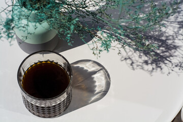 Faceted glass with black filter coffee on arabika beans in third-wave coffee shop, on the background of white table with blue flowers.