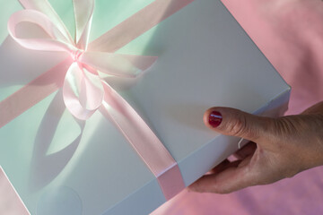 White gift box with a gentle pink bow in female hands on a pink background. Acceptance of a gift for the holiday. High quality photo