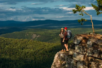Foto op Aluminium A man and a woman in tourist equipment are standing on a rock and admiring the panoramic view. Two tourists with backpacks enjoy the sunset view from the top of the mountain. Backpacking trip © MISHA