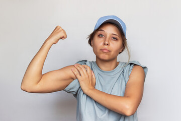 Strong confident caucasian young blonde woman in a gray t-shirt and cap raises arm and shows bicep...