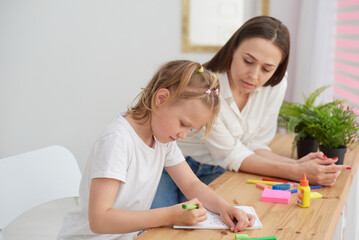 Obraz na płótnie Canvas Mom and daughter are doing homework. A little girl thoughtfully completes math assignments under the supervision of a tutor. The concept of taking care of a child and helping with homework