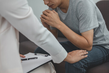Asian young man, male suffers from a mental who needs to therapy with a psychologist while sitting on couch to consult, psychiatrist has encouragement the patient by touching to make his feel relaxed.