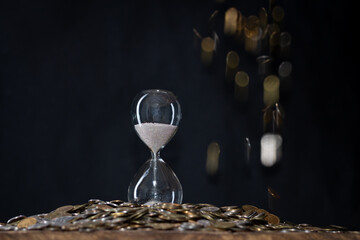 A composition with an hourglass standing on silver and yellow coins lying on a table and falling on a dark background. Selective focus. Concept: time is money