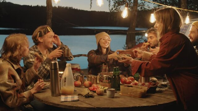 Company of young friends sitting together at table under garland, sharing dishes and eating food while having dinner at campsite in evening