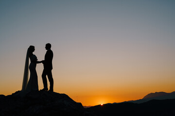 Silhouettes of the bride and groom stand on the mountain against the background of the sunset