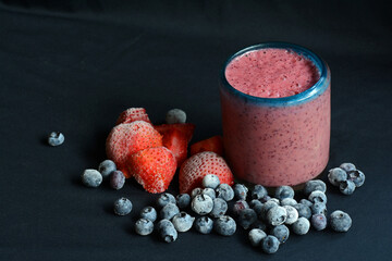 Frozen blueberries and strawberries on table in front of  smoothie glass with ingredients on black background.