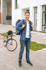 Full length vertical portrait of joyful handsome young delivery man with thermo backpack standing on city street, on background of bicycle and office building, looking at camera.