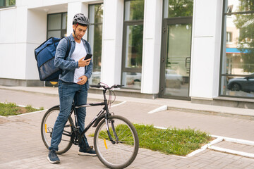 Fototapeta premium Medium shot of handsome male courier with thermo backpack standing with bicycle in city street and using navigation app on phone. Delivery man looking for client address looking smartphone.
