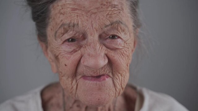Portrait of Caucasian toothless senior woman 90 years old with deep wrinkles and gray hair cute smiling and talking on gray background in studio. Old female with dental problems. Orthodontist patient.