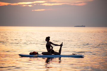 a female makes yoga asana on the SUP board at sea sunrise