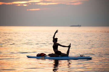 a female makes yoga asana on the SUP board at sea sunrise