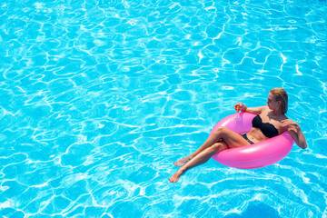 A young woman swims on a pink inflatable circle in clear blue water. A girl with a cocktail relaxes...