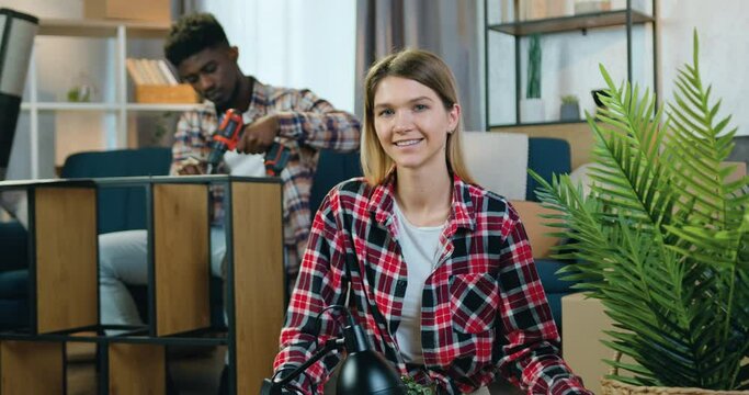 Close up of beautiful happy joyful blond girl which looking into camera with sincerely smile on the background of her black-skinned male friend which assembling rack with electric screwdriver