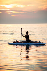 a female makes yoga asana on the SUP board at the orange light of sunrise