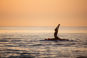 a female makes yoga asana on the SUP board at the orange light of sunrise