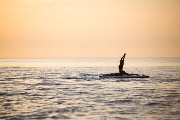 a female makes yoga asana on the SUP board at the orange light of sunrise
