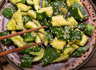 Broken cucumbers in a plate sprinkled with sesame seeds and chopsticks hold a piece.