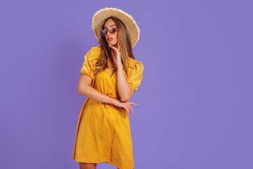 Portrait of excited young woman in yellow dress, summer hat, sunglasses posing isolated on pastel violet background.