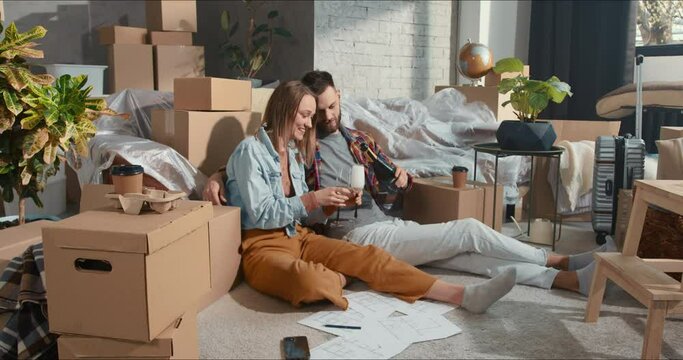 Celebrating new home. Young happy couple enjoying wine relaxing after unpacking in apartment with stuff in removal boxes