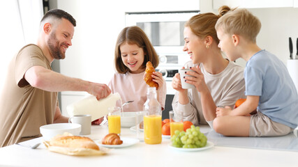 Obraz na płótnie Canvas Young happy family with two cute little kids having breakfast together in kitchen and smiling