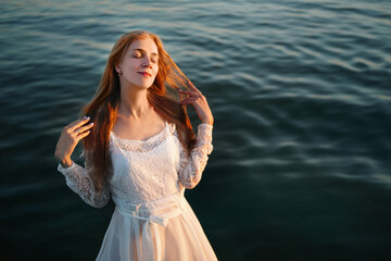 Charming woman is resting at the sea. Clear ocean water, beautiful reflection on waves. White wedding dress, lace sleeves, long orange hair. Yellow sunset leaves shadows on a happy face. Closed eyes