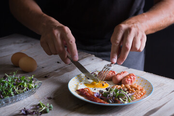 Man's hands are cutting a fried egg on a plate. English breakfast, eggs, sausages, bacon and beans