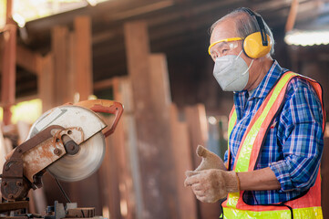 Asian elderly carpenter craftsman in carpentry shop use circular saws to cut wood board make furniture, wear safety equipment at work such as dust masks, glasses, gloves, noise-canceling, headphones.