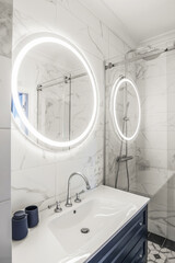 Modern bathroom interior with a dark blue wooden shelf, white sink and big round mirror with LED light. - obrazy, fototapety, plakaty