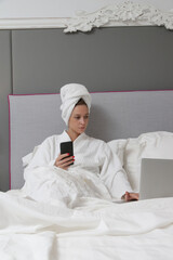Young caucasian woman using cellphone and laptop in bed 
