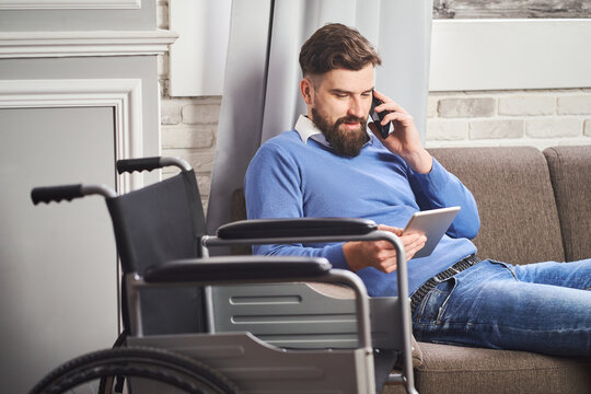 Disabled man sitting on a couch, using a tablet computer and talking on a phone