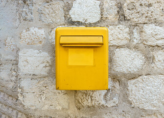 A yellow mailbox on a light stone wall. Box for letters and postcards.