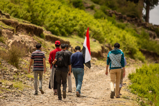 A group of Yemeni youth walking on a dirt road surrounded by trees and nature, carrying the Yemeni flag. Yemen traveler.