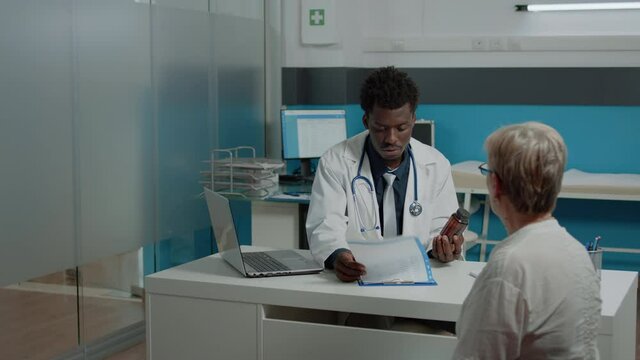 Adult doctor of african ethnicity giving bottle of pills to sick aged patient in medical office. Young black medic holding medicine, treatment or drugs for healing disease for healthcare