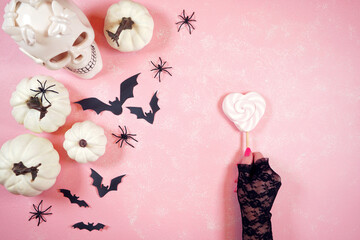 Modern pink Halloween theme flatly background with woman's hand holding lollipop, white skull, pumpkins, lollipops and bats.