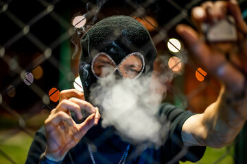 young man in balaclava looking at camera and blow smoke out. crime and violence, drugs, fashion, rap, rapper new school, style in night city. - obrazy, fototapety, plakaty
