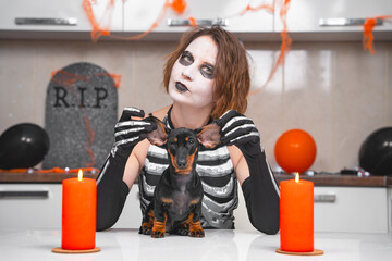 Young sullen woman with scary makeup in skeleton costume examines dachshund puppy on table,...
